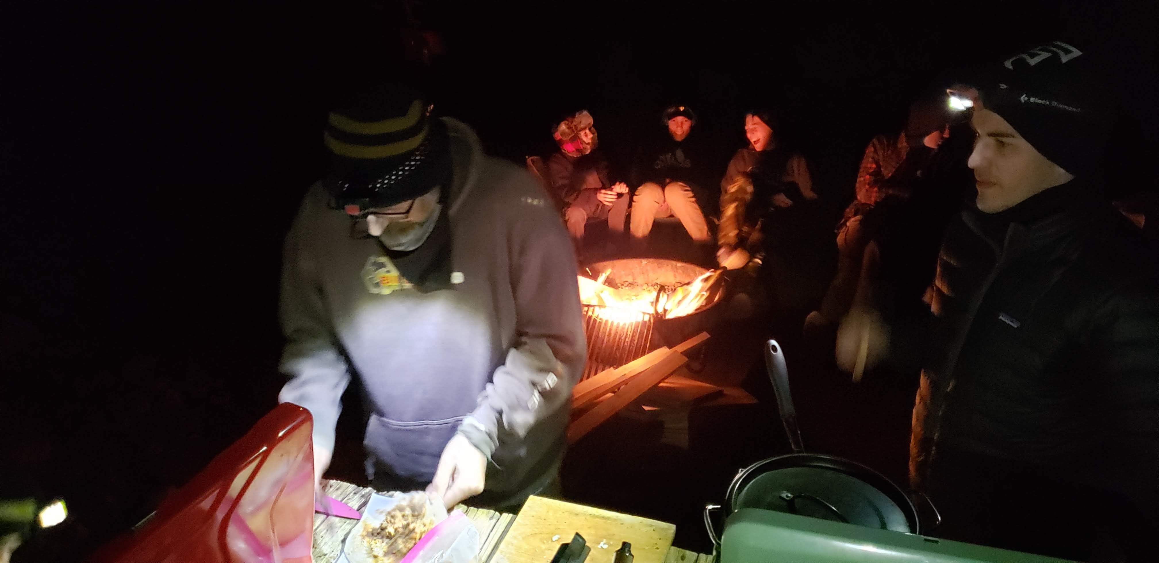 Boys around a campfire while men prepare burritos for dinner.