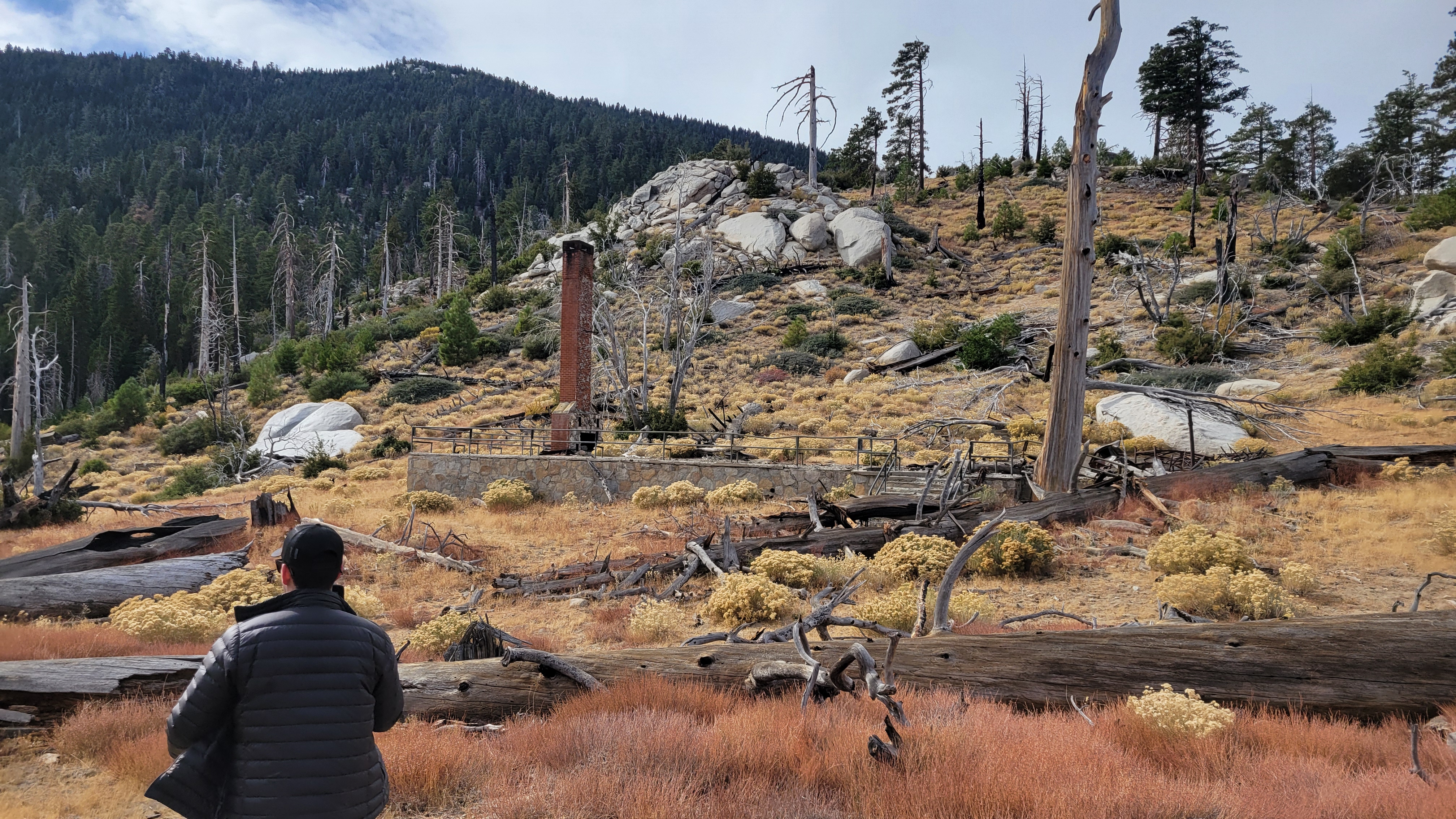 A person walking towards ruins of a stone foundation and red brick chimney.