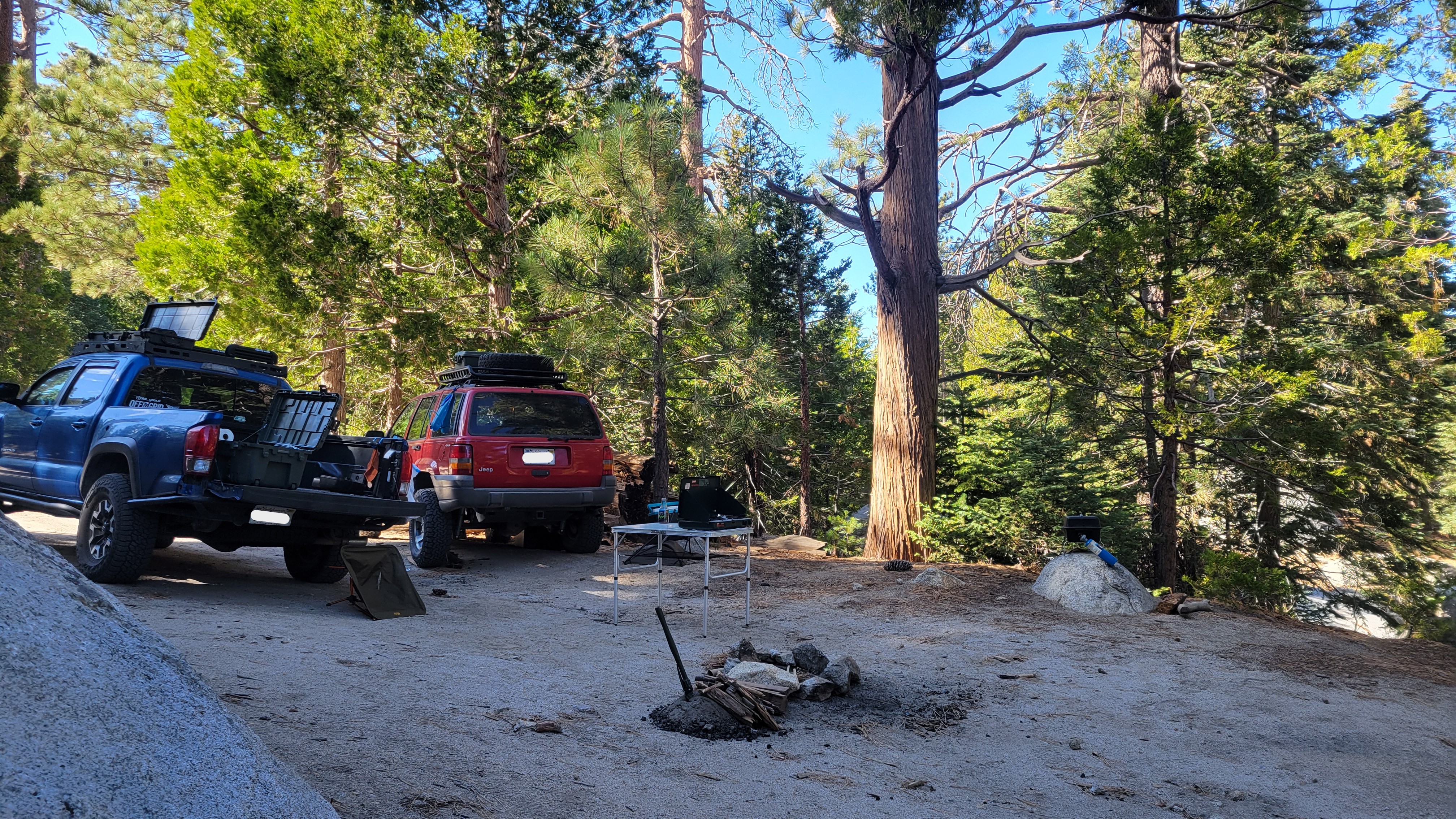 A blue Tacoma and a red Jeep make camp in a clear, flat area. A fire pit and folding table make up the center of the scene.